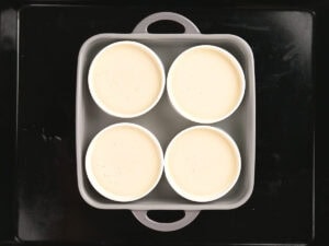 Four white ramekins filled with a pale liquid are arranged in a square baking dish, placed on a dark baking tray.