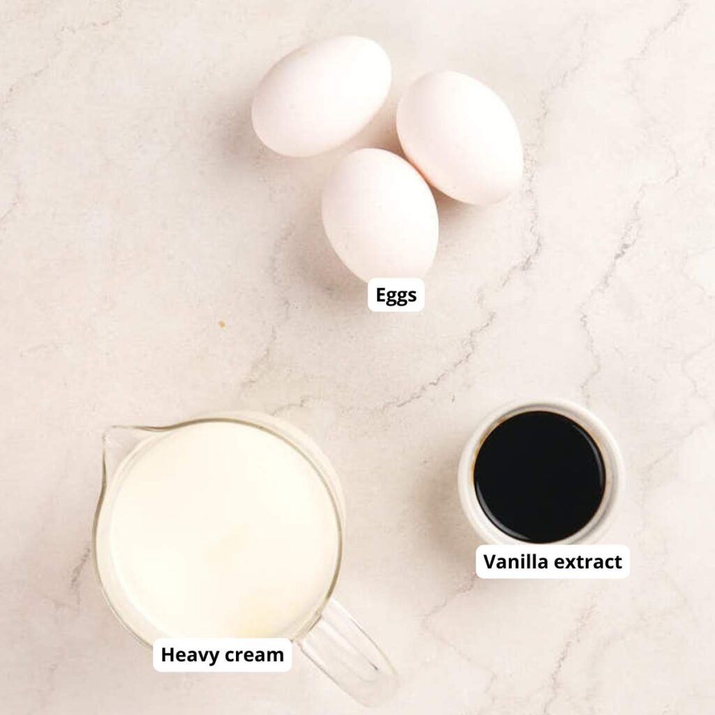 Three eggs, a measuring cup of heavy cream, and a small bowl of vanilla extract are arranged on a light marble surface. Each item is labeled.