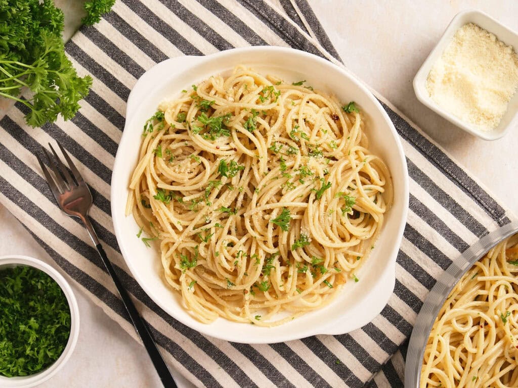 A bowl of cooked spaghetti topped with chopped parsley and grated cheese, placed on a striped cloth with a fork and parsley garnish nearby.