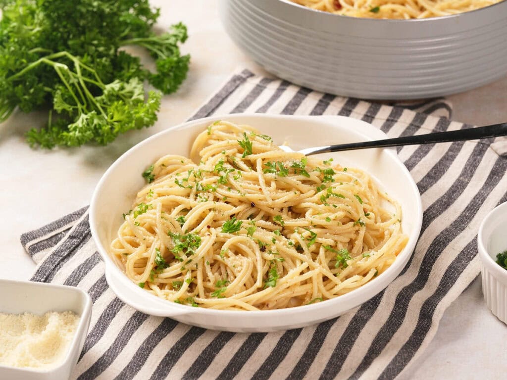 A bowl of spaghetti garnished with chopped parsley sits on a striped cloth next to a fork, grated cheese, and fresh herbs.