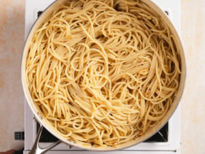 A pot of plain cooked spaghetti sits on a stovetop burner, lightly coated with oil and small bits of seasoning visible.