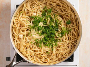 A pan of cooked spaghetti topped with chopped parsley, grated cheese, and minced garlic sits on a stove burner.
