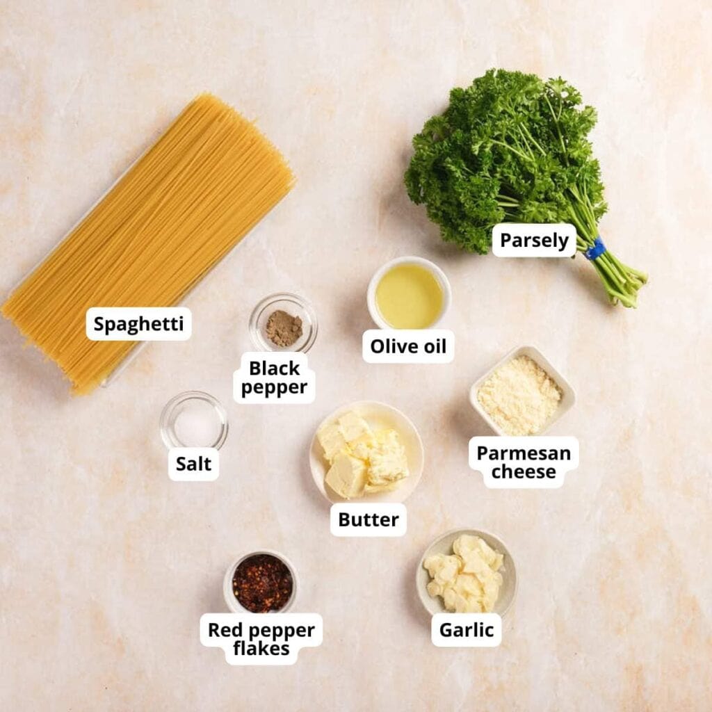 Ingredients for spaghetti include uncooked pasta, parsley, olive oil, black pepper, salt, parmesan cheese, butter, garlic, and red pepper flakes arranged on a light surface.