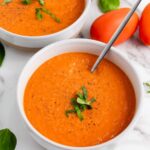 Two white bowls of tomato soup garnished with chopped basil sit on a marble surface, each with a spoon, surrounded by fresh basil leaves and whole tomatoes.