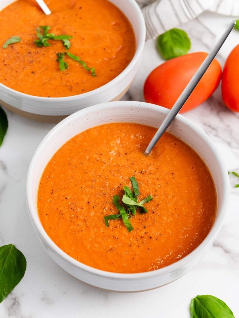 Two white bowls of tomato soup garnished with chopped basil sit on a marble surface, each with a spoon, surrounded by fresh basil leaves and whole tomatoes.