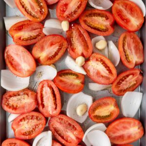 Halved tomatoes, quartered onions, and whole garlic cloves arranged on a baking tray, seasoned with salt and pepper, ready for roasting.