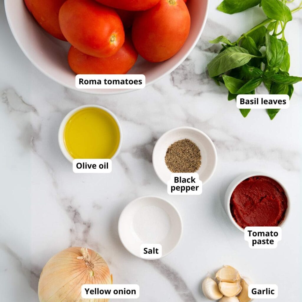 Top-down view of Roma tomatoes, basil leaves, olive oil, black pepper, tomato paste, salt, yellow onion, and garlic arranged on a marble surface, each labeled.