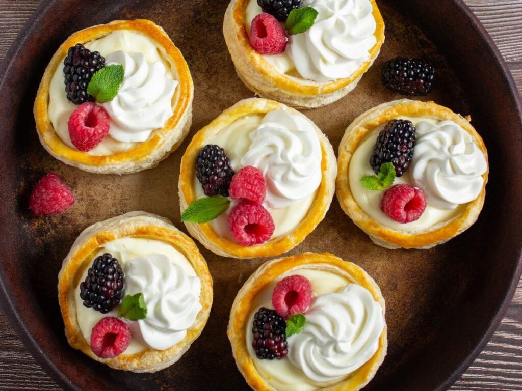 Six mini cheesecakes topped with whipped cream, raspberries, blackberries, and mint leaves are arranged on a round dark plate.