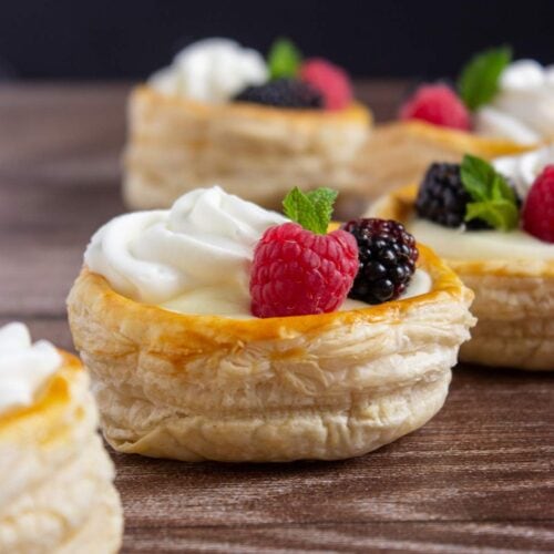 Round puff pastries filled with cream, topped with raspberries, blackberries, and mint leaves, arranged on a wooden surface.