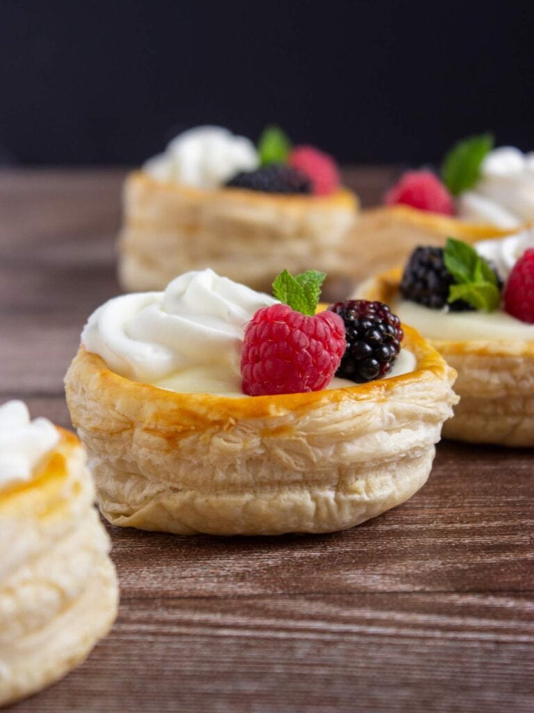 Round puff pastries filled with cream, topped with raspberries, blackberries, and mint leaves, arranged on a wooden surface.