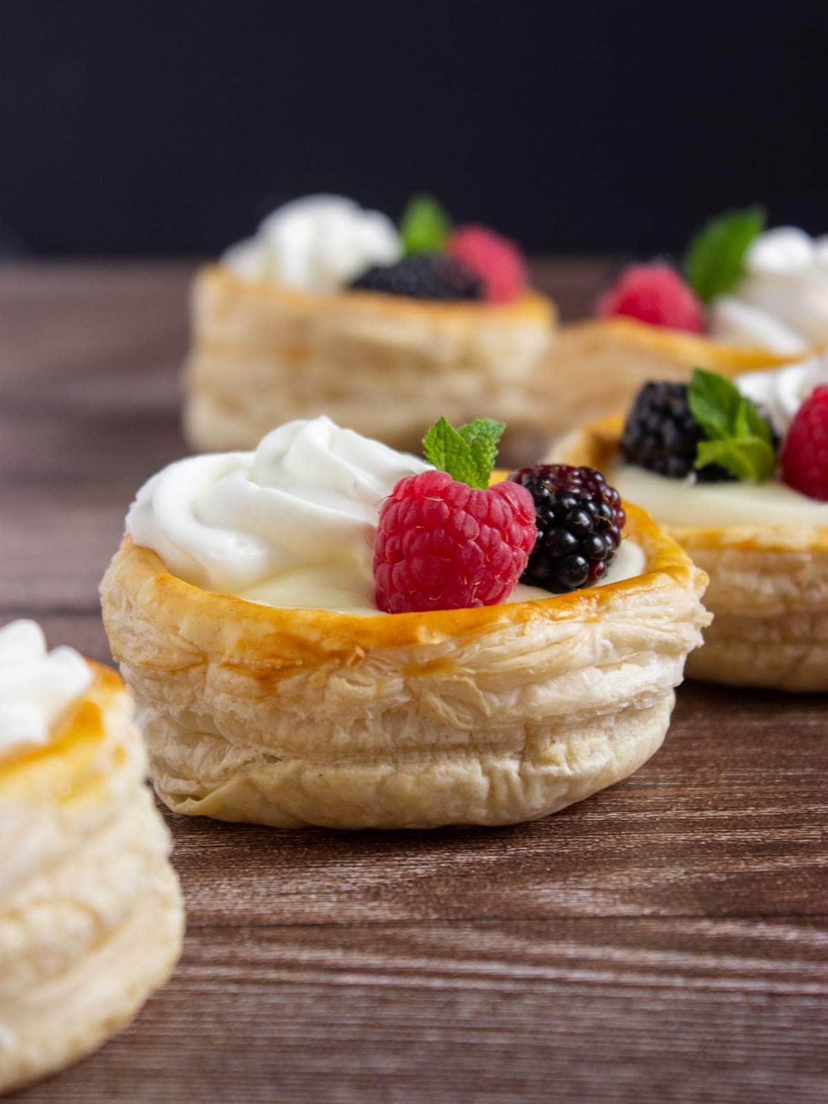 Round puff pastries filled with cream, topped with raspberries, blackberries, and mint leaves, arranged on a wooden surface.