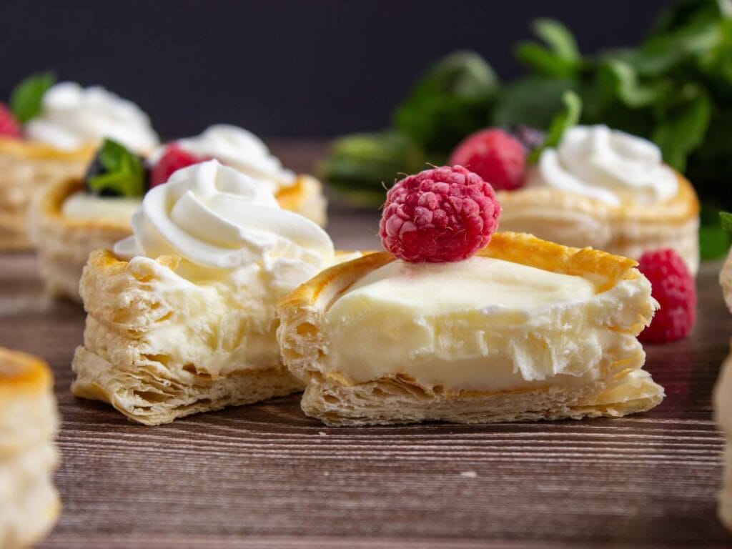A close-up of a flaky puff pastry dessert filled with cream, topped with whipped cream and a raspberry, with more pastries and raspberries in the background.