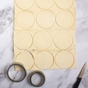 Rectangular sheet of dough with circular shapes cut out, two metal cutters, and a knife placed on a marble surface.