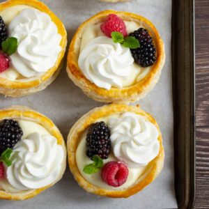 Four puff pastry tartlets filled with cream, topped with whipped cream, blackberries, raspberries, and mint leaves, arranged on a parchment-lined baking tray.