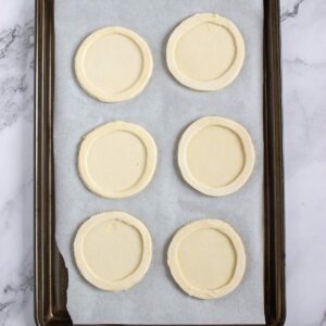 A baking sheet lined with parchment paper holds six unbaked round pastry dough pieces, each with an inner circle indentation.