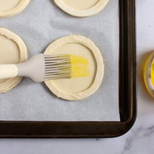 A pastry brush applies egg wash to round puff pastry dough on a baking sheet lined with parchment paper. A bowl of egg wash is visible beside the tray.