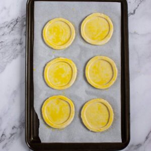 A baking tray lined with parchment paper holds six round, uncooked pastry circles brushed with egg wash.