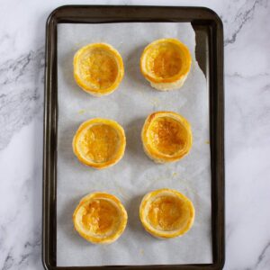 Six empty golden-brown pastry shells on a parchment-lined baking tray, placed on a marble surface.