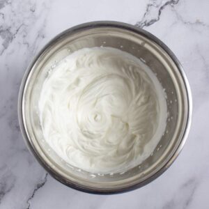 A stainless steel bowl filled with freshly whipped cream sits on a white marble surface.