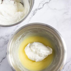 Two metal bowls on a marble surface; one with whipped cream and the other with a mixture of whipped cream and yellow batter being combined.