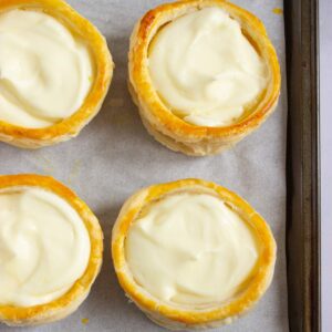 Four round pastry shells filled with creamy white custard, arranged on parchment paper on a baking tray.