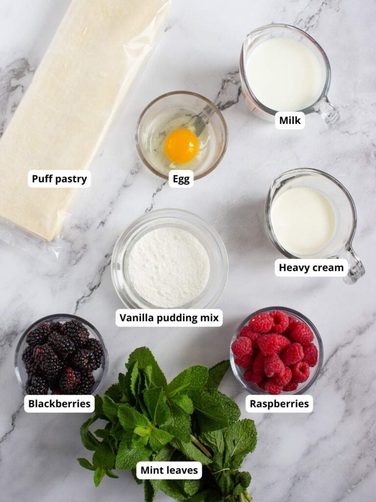 Ingredients on a marble surface, including puff pastry, egg, milk, heavy cream, vanilla pudding mix, blackberries, raspberries, and mint leaves, each labeled.