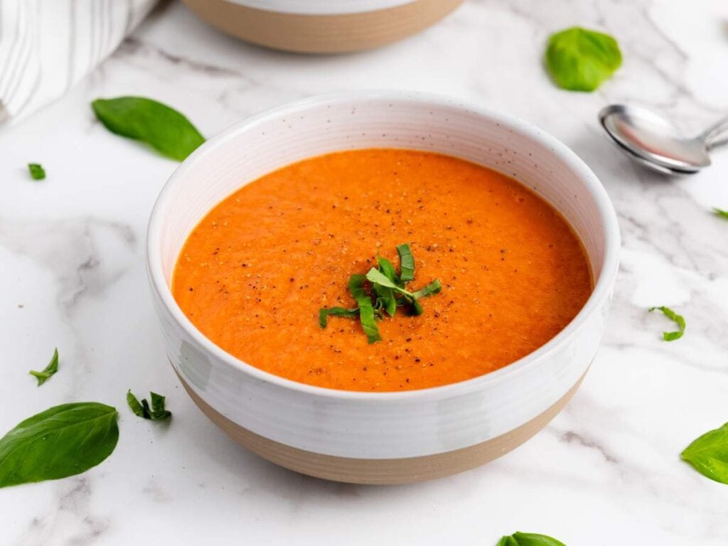 A bowl of tomato soup garnished with chopped basil, with fresh basil leaves and a spoon on a marble surface.