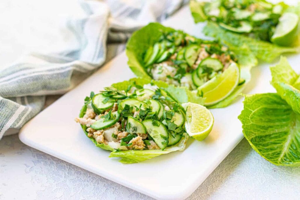 Lettuce wraps filled with ground chickem, sliced cucumber, herbs, and lime wedges on a white rectangular plate.